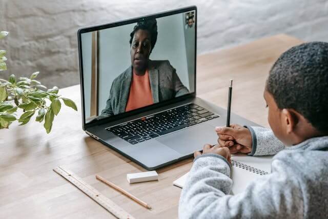 Clever black boy taking notes and listening to teacher online